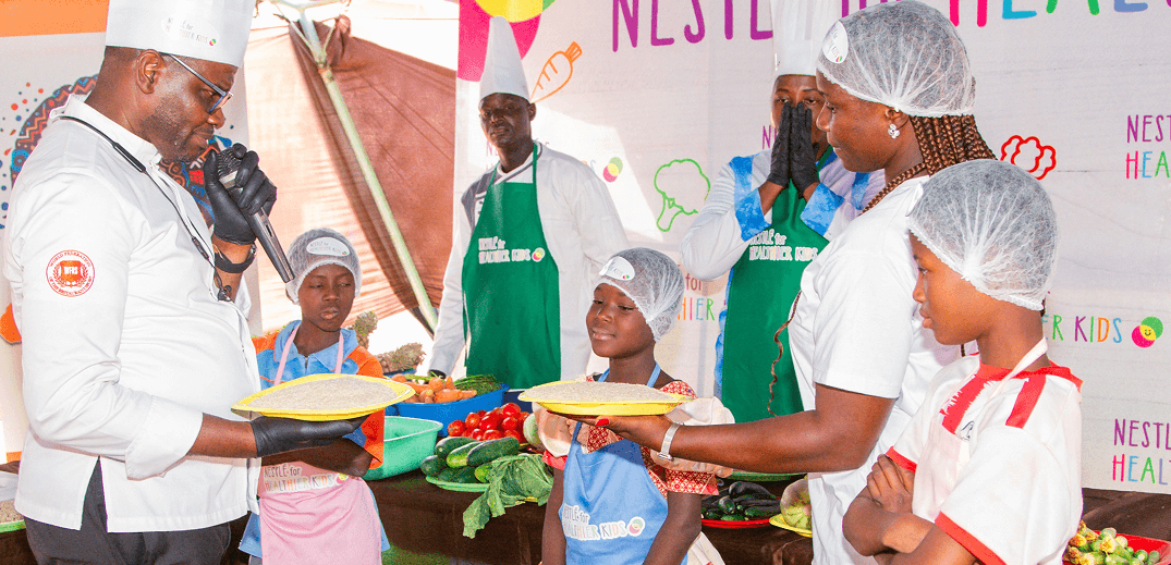 A chef presents a colorful dish to a group of children wearing aprons and chef hats, surrounded by fresh vegetables
