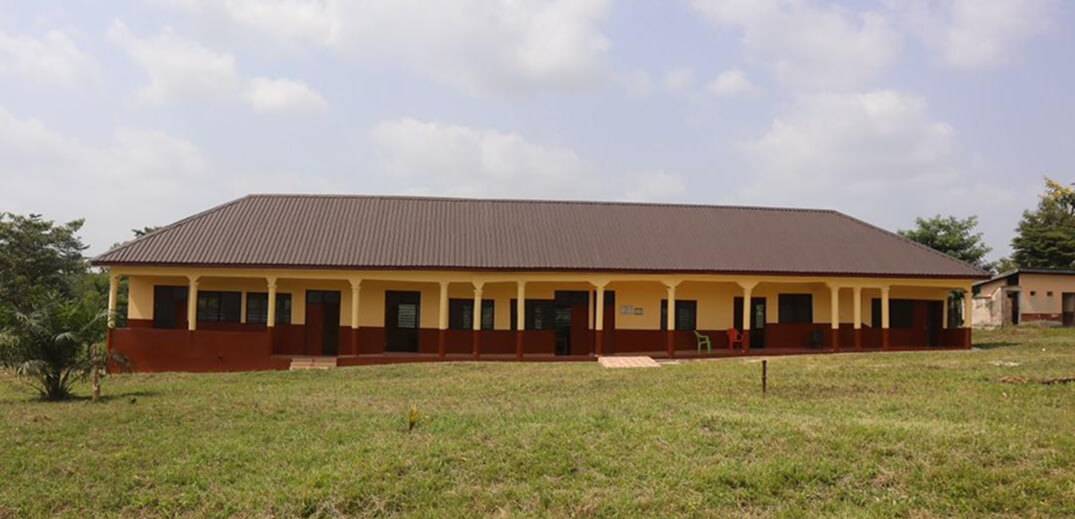A single-story school building with a yellow and brown exterior, surrounded by greenery under a partly cloudy sky.