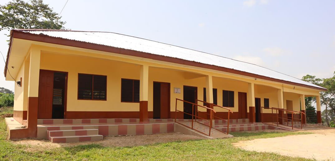 A single-story school building with a yellow and brown exterior, surrounded by greenery under a partly cloudy sky.