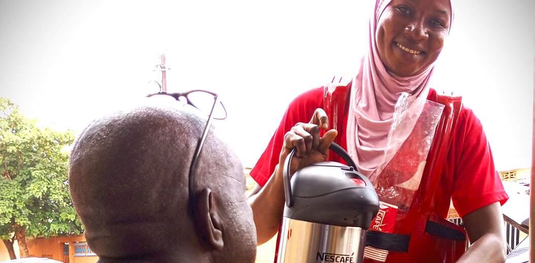 A person in a red shirt and hijab holds a thermos labeled "Nescafé," while serving an elderly individual outdoors.