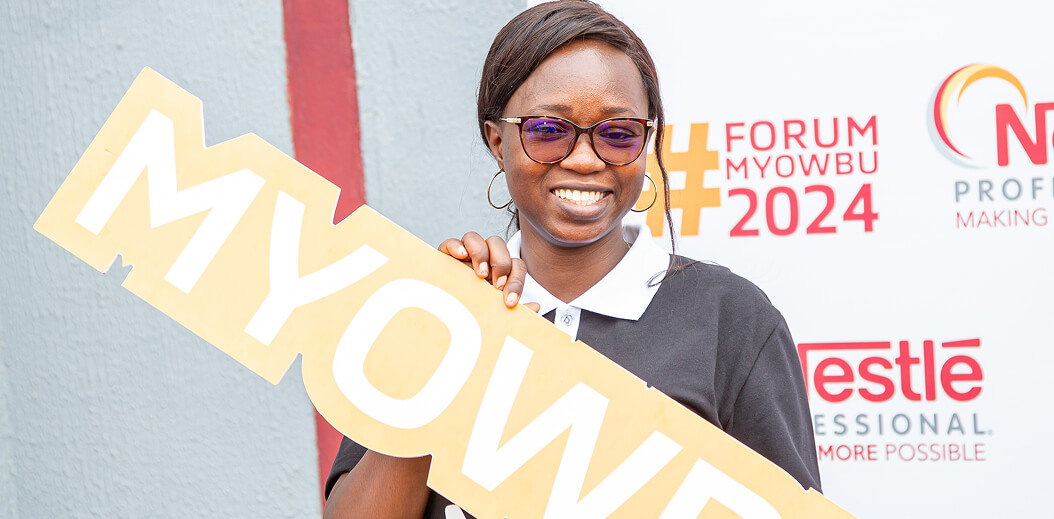 A person stands against a colorful backdrop, holding a large sign that reads "MYOWBU," promoting the #FORUMMYOWBU2024 event.