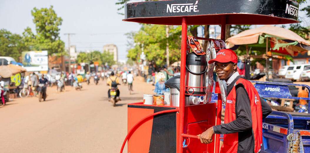 A street vendor serves Nescafe from a bright red cart, with a busy road and shoppers in the background under a clear blue sky