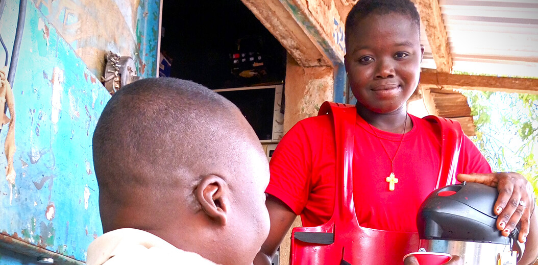 A vendor in a red shirt serves tea from a kettle to a customer outside a rustic shop with peeling paint and warm sunlight.