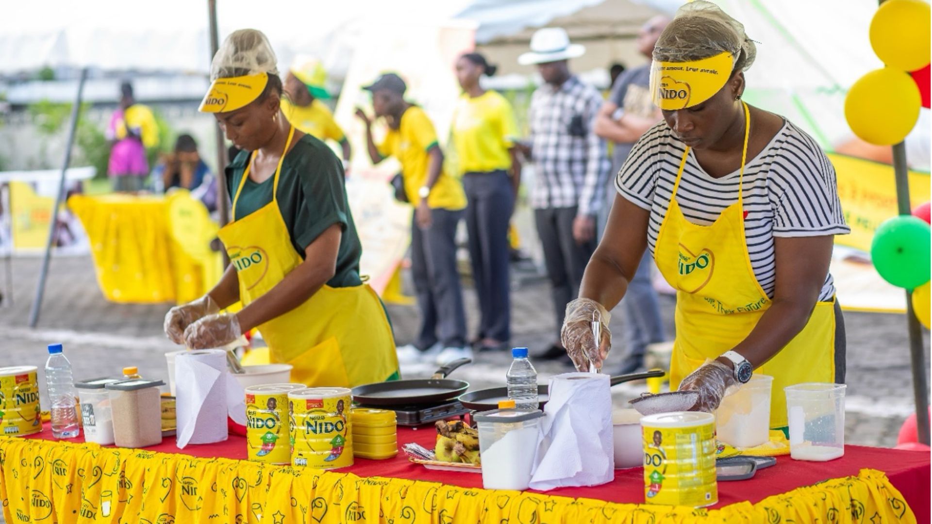 Two women wearing yellow Nido aprons and gloves prepare food at a colorful booth with Nido products and cooking supplies.