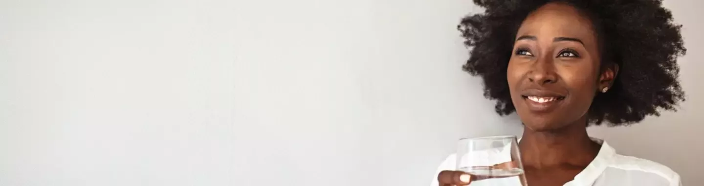 Smiling woman with natural hair holding a glass of water against a light background.
