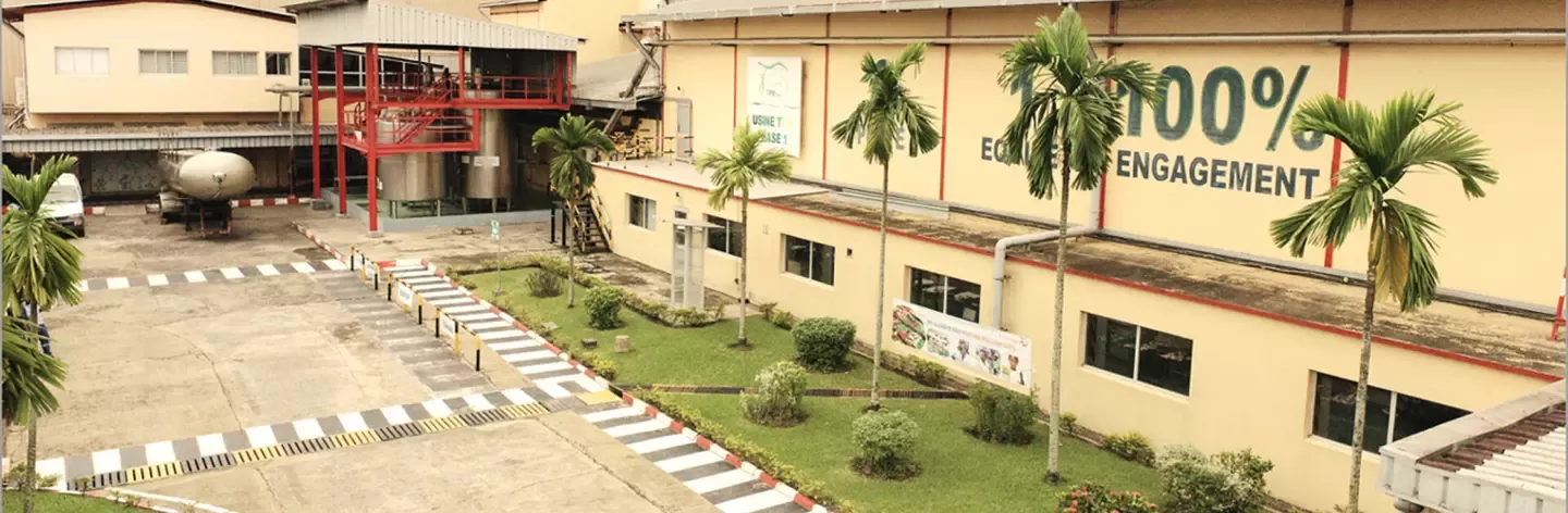 A factory building featuring greenery, palm trees, and a prominent banner stating "100% ENGAGEMENT" on its side, showcasing an industrial setting.