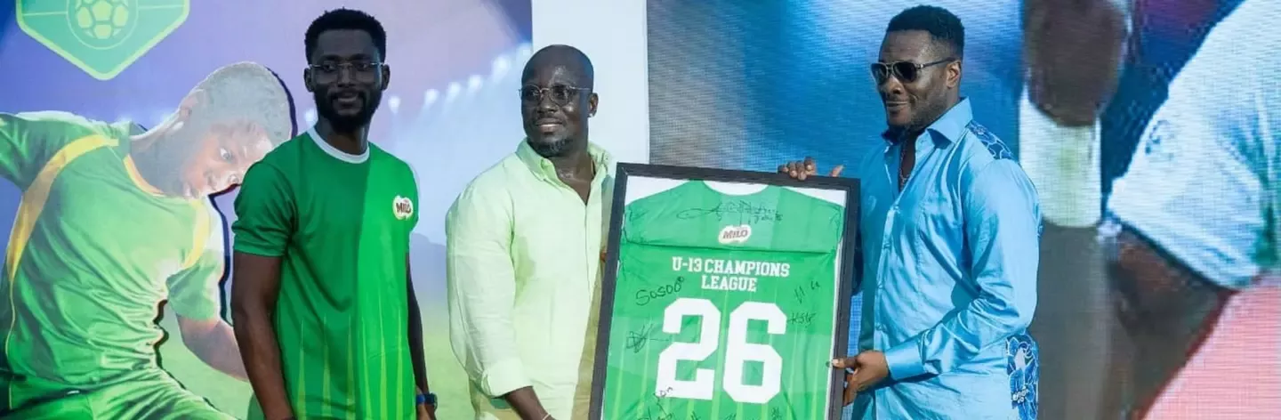 Three men presenting a framed green sports jersey with "U-13 Champions League 26" and signatures on it.