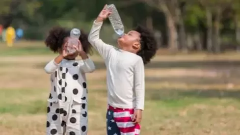 Two children playfully drink from water bottles outdoors in a park, with one wearing a polka-dot costume and the other American flag shorts.