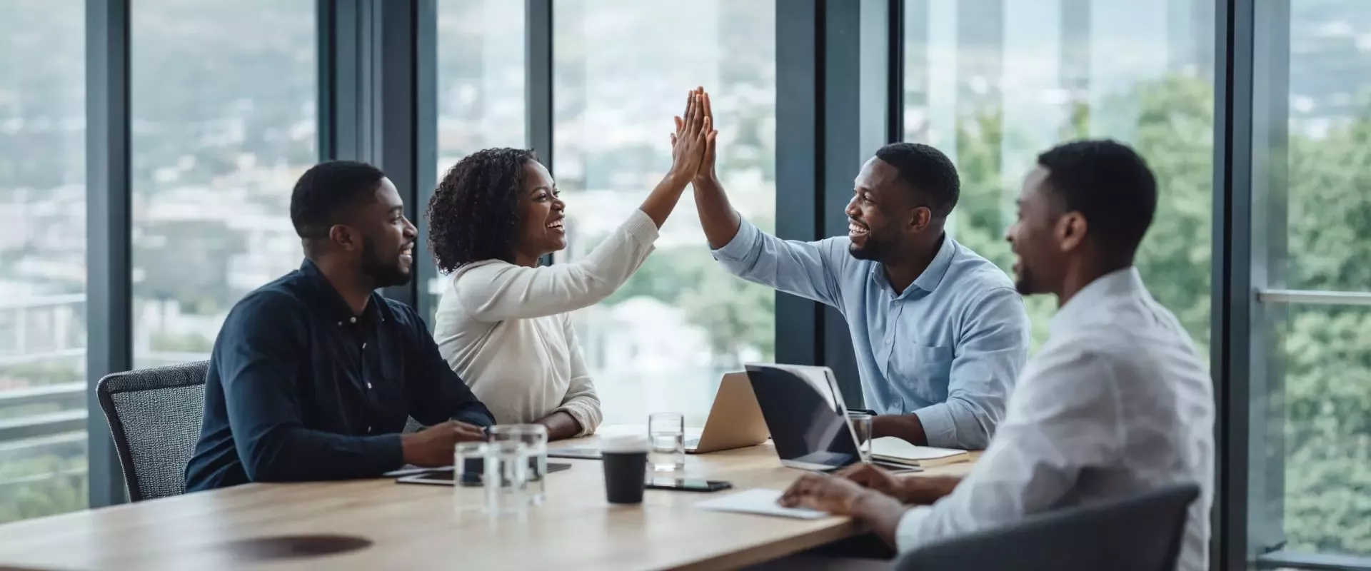 Diverse business team in meeting room giving a high-five, collaborating with laptops and documents on table, city views outside.