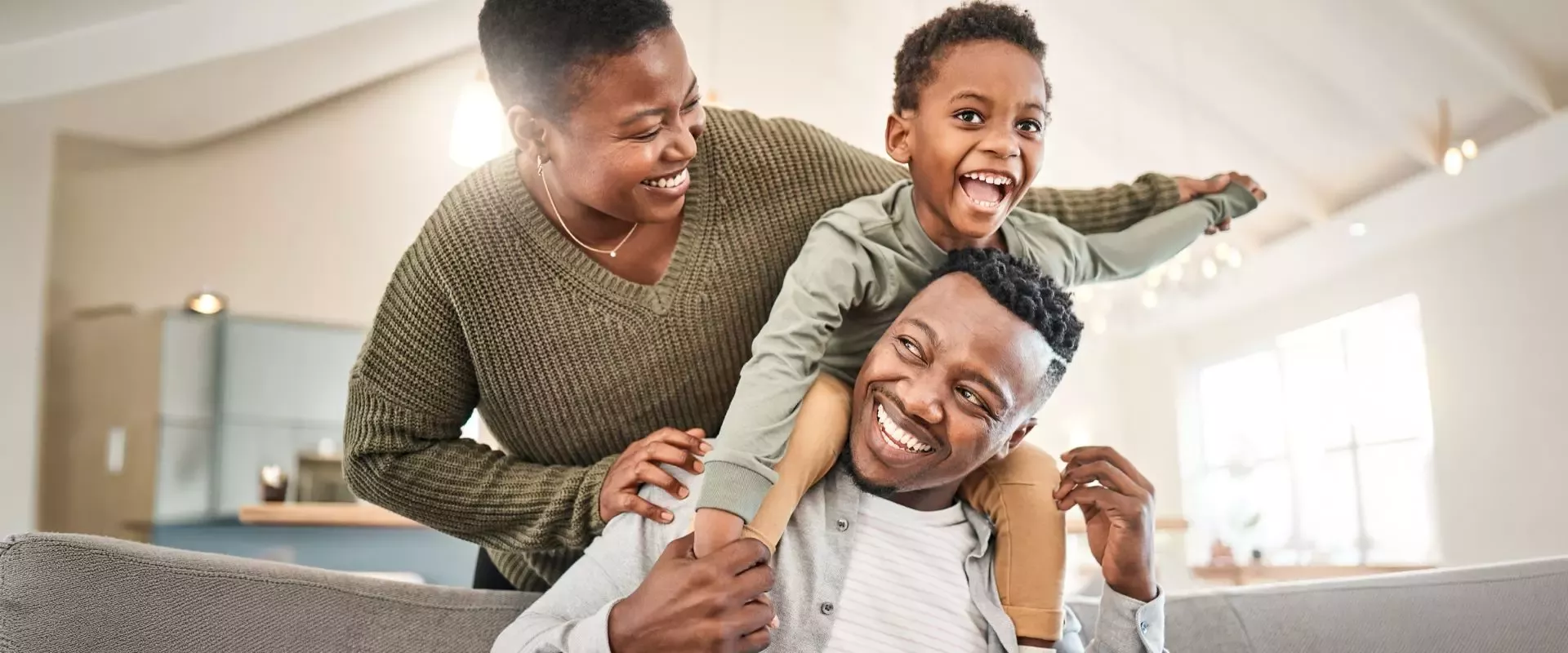 Three people having fun indoors; a child sits on a man's shoulders while a woman supports them and smiles.