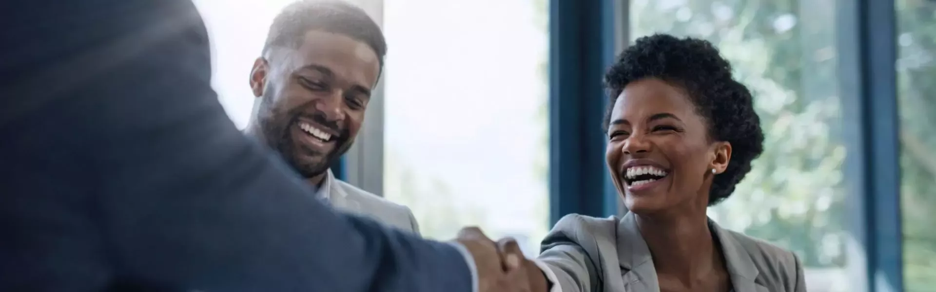Two business professionals shaking hands in a bright office with large windows and a blurred background.