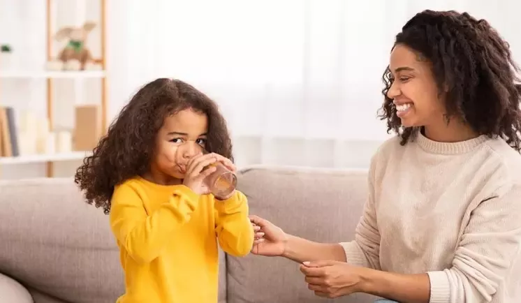 Smiling mother watches her young daughter drink from a glass while sitting together on a sofa.