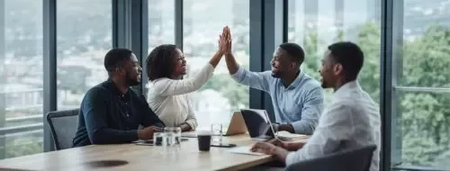 Diverse business team in meeting room giving a high-five, collaborating with laptops and documents on table, city views outside.