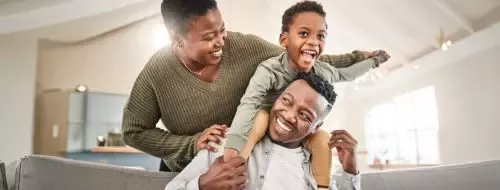 Three people having fun indoors; a child sits on a man's shoulders while a woman supports them and smiles.