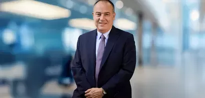 A professionally dressed man stands confidently with arms crossed, set against a softly blurred modern office background.