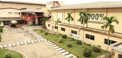 A factory building featuring greenery, palm trees, and a prominent banner stating "100% ENGAGEMENT" on its side, showcasing an industrial setting.