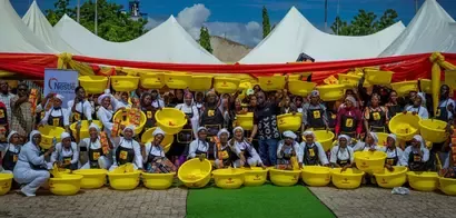 A group of individuals holding yellow bowls and food products, gathered in front of a festive backdrop with tents and decorations