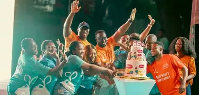 A lively group of people enthusiastically reaches toward a decorated cake topped with colorful products and a prominent "85%" sign.