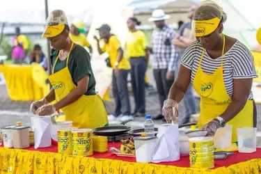 Two women wearing yellow Nido aprons and gloves prepare food at a colorful booth with Nido products and cooking supplies.