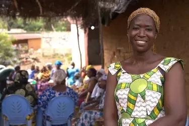 Smiling woman in a colorful patterned dress and gold headpiece stands before a rural gathering seated under a thatched shelter.