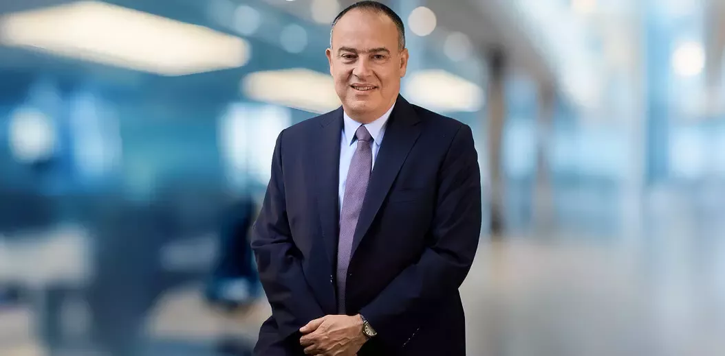A professionally dressed man stands confidently with arms crossed, set against a softly blurred modern office background.