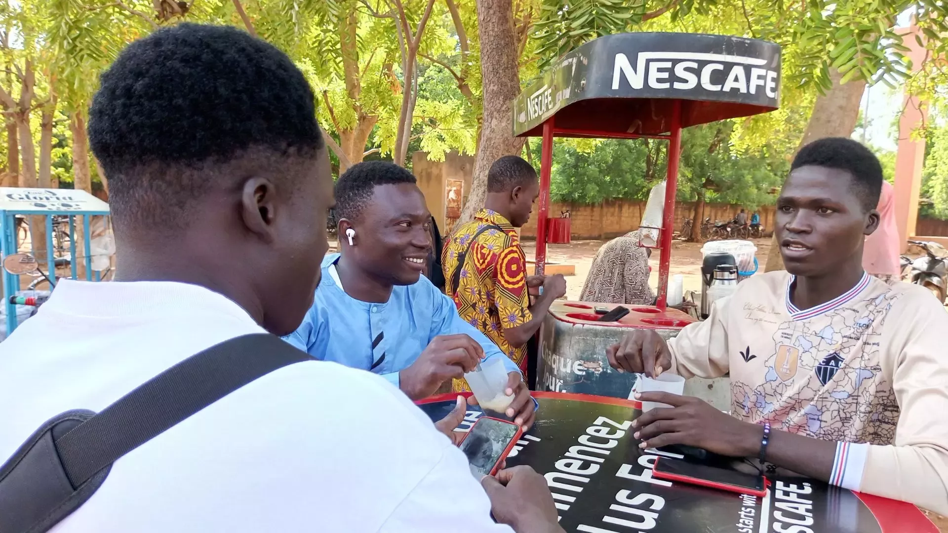 A group of young men gather around a table at a street kiosk, discussing while enjoying beverages under lush greenery.