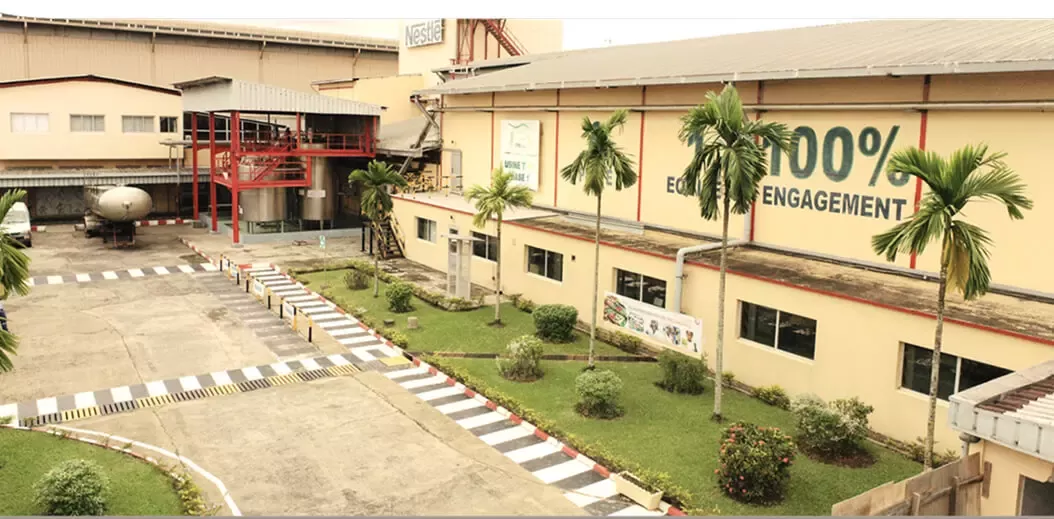 A factory building featuring greenery, palm trees, and a prominent banner stating "100% ENGAGEMENT" on its side, showcasing an industrial setting.