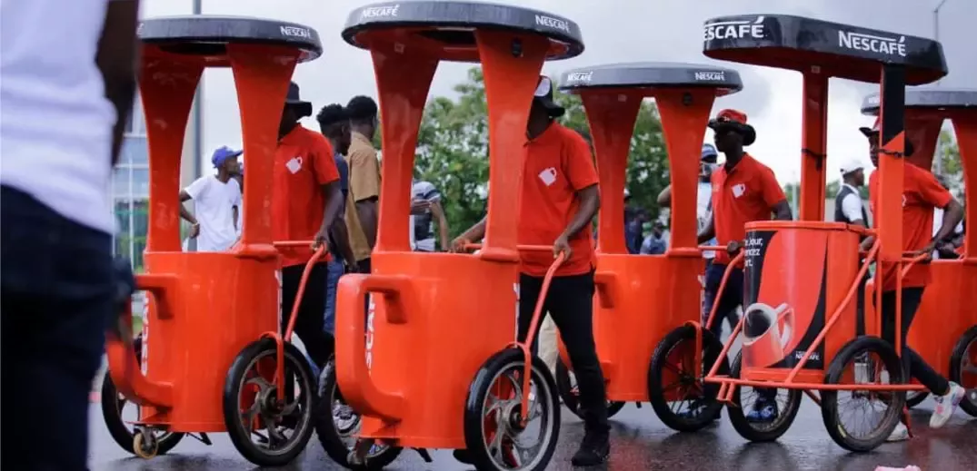 A group of vendors in red shirts push orange coffee carts with NESCAFÉ branding along a street during an event.