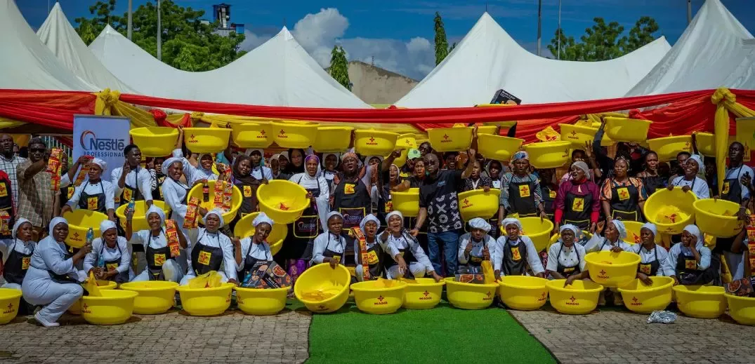 A group of individuals holding yellow bowls and food products, gathered in front of a festive backdrop with tents and decorations