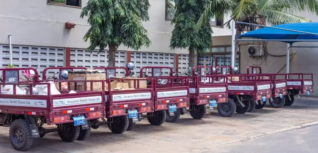 A row of red, utility trucks loaded with goods, marked as donations from Nestlé Ghana Ltd., beside green trees in a courtyard.