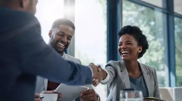 Two business professionals shaking hands in a bright office with large windows and a blurred background.