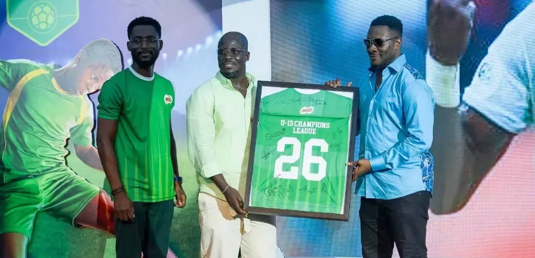 Three men presenting a framed green sports jersey with "U-13 Champions League 26" and signatures on it.