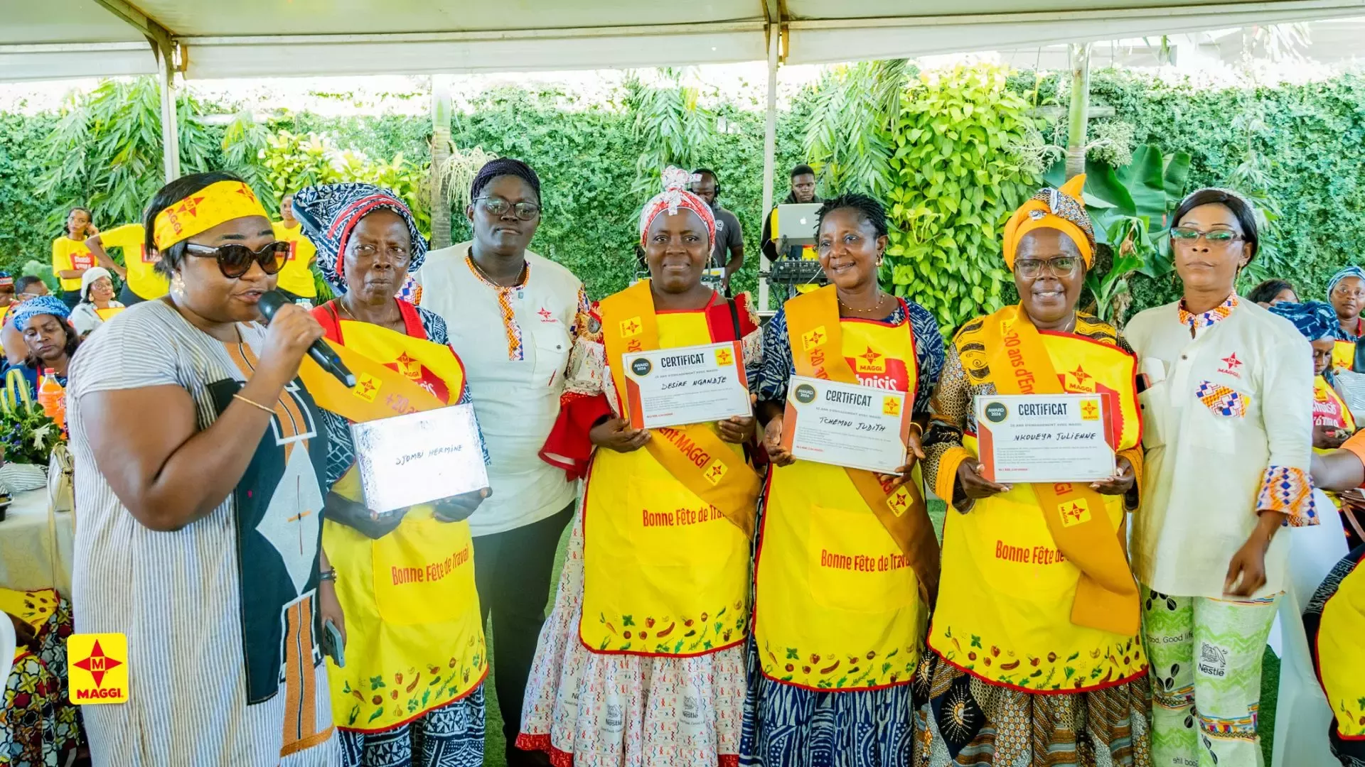 Group of women wearing yellow Maggi aprons and sashes, holding certificates at an outdoor event celebrating Labor Day.