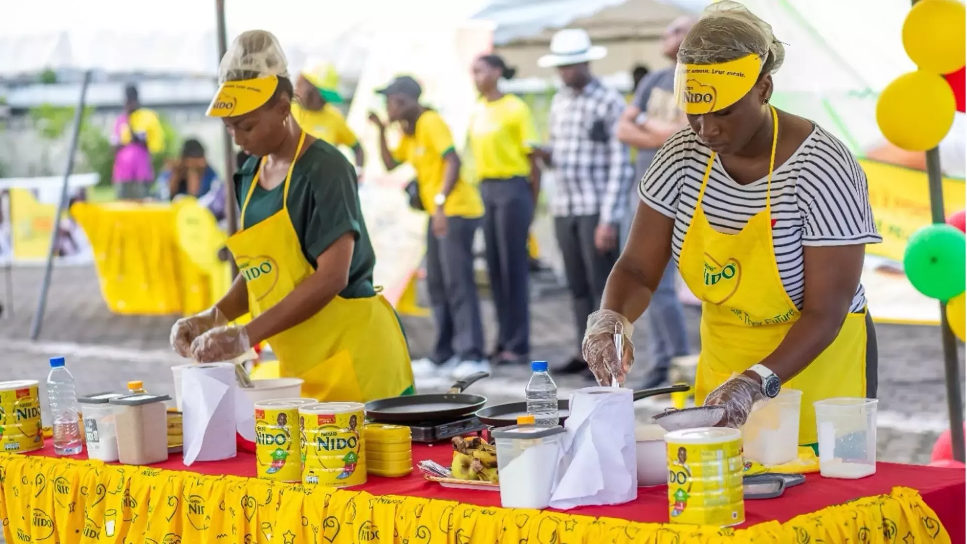 Two women wearing yellow Nido aprons and gloves prepare food at a colorful booth with Nido products and cooking supplies.