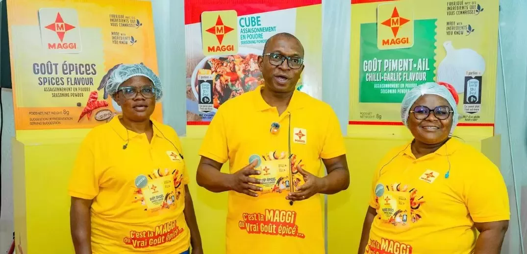 Three people in yellow Maggi shirts and hairnets pose in front of large Maggi seasoning powder ads in spices, cube, and chili-garlic flavors.