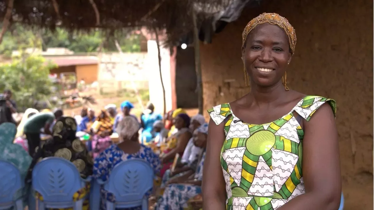 Smiling woman in a colorful patterned dress and gold headpiece stands before a rural gathering seated under a thatched shelter.